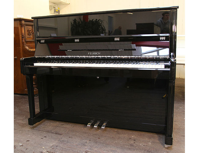 A brand new, Feurich Model 122 upright piano with a black case and chrome fittings. Piano has an eighty-eight note keyboard and three pedals.
